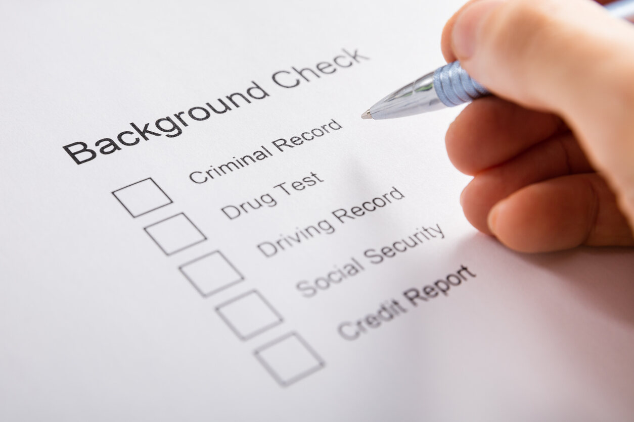 Hand with a pen checking boxes on a "Background Check" form for criminal, drug, driving, and credit records, relevant to citizenship applications.